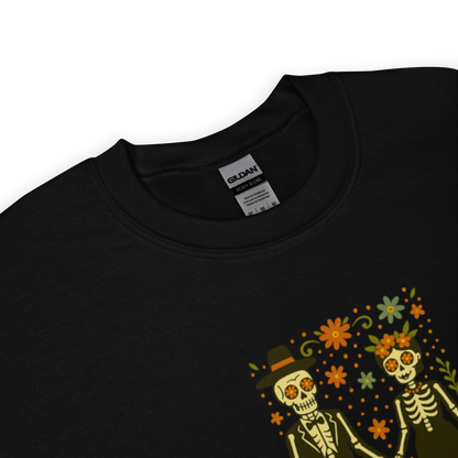 This is a zoom in on a black sweatshirt with a Día de los Muertos–style illustration of a male and female skeleton holding hands, surrounded by marigolds, vines, and folk art patterns. The skeletons wear traditional black clothing, the man in a suit and hat, the woman with a floral crown with a red heart and leaves at the bottom and the text Creepy All Year. Rendered in warm tones of orange, green, and cream, symbolizing eternal love and remembrance