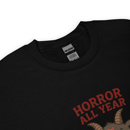 This is a zoom in of a black sweatshirt with a cartoon illustration of a baby Baphomet goat with curved horns and a playful grin, wearing bright floral swim trunks while floating in a colorful inner tube. The character is set against a light background with the bold text “Horror All Year” above in red letters
