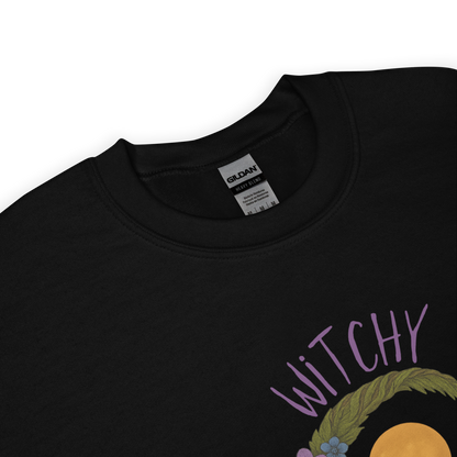This is a zoom in of a design on a black sweatshirt of a hand-drawn wreath made of moss-covered branches wraps around a glowing sun. The wreath is decorated with purple and blue flowers, small amethyst and fluorite crystals, and strands of hanging moss. The words “Witchy” curve above the wreath and “All Year” sits on the bottom right