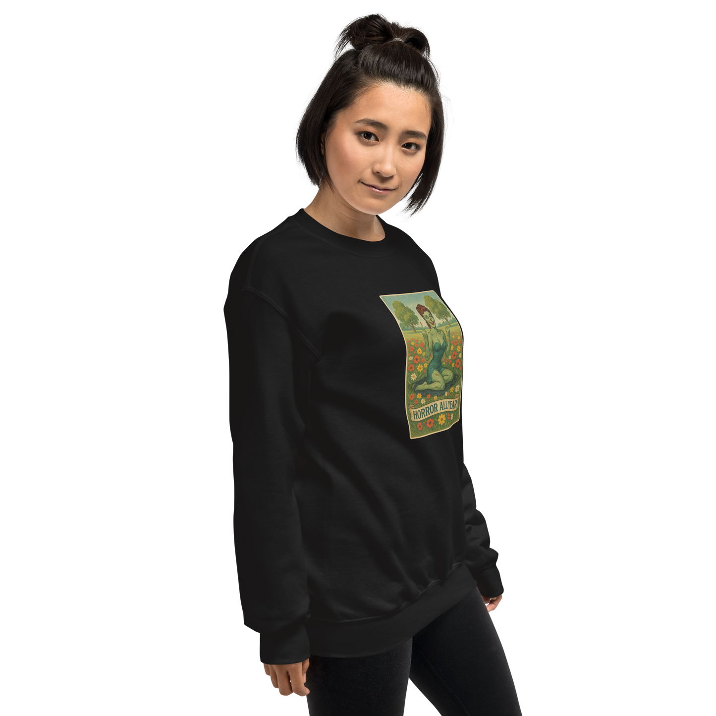 This is a woman with dark hair wearing a black sweatshirt with a design illustration of a sassy zombie pinup girl standing in a floral field, wearing tattered lingerie and flipping off the viewer with both hands. She has green skin, vintage curls, and a bold attitude. The design blends horror and humor, with a pastel color palette and creepy cute vibes