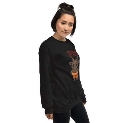 This is a woman with dark hair wearing a black sweatshirt with a cartoon illustration of a baby Baphomet goat with curved horns and a playful grin, wearing bright floral swim trunks while floating in a colorful inner tube. The character is set against a light background with the bold text “Horror All Year” above in red letters
