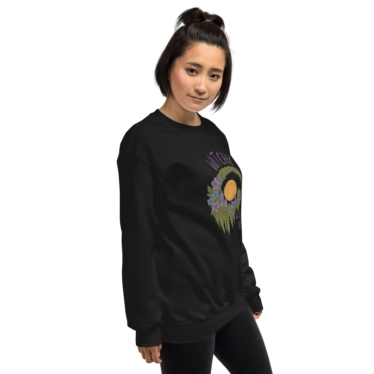 This is a woman with dark hair wearing a black sweatshirt with a design of a hand-drawn wreath made of moss-covered branches wraps around a glowing sun. The wreath is decorated with purple and blue flowers, small amethyst and fluorite crystals, and strands of hanging moss. The words “Witchy” curve above the wreath and “All Year” sits on the bottom right 