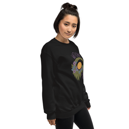 This is a woman with dark hair wearing a black sweatshirt with a design of a hand-drawn wreath made of moss-covered branches wraps around a glowing sun. The wreath is decorated with purple and blue flowers, small amethyst and fluorite crystals, and strands of hanging moss. The words “Witchy” curve above the wreath and “All Year” sits on the bottom right 