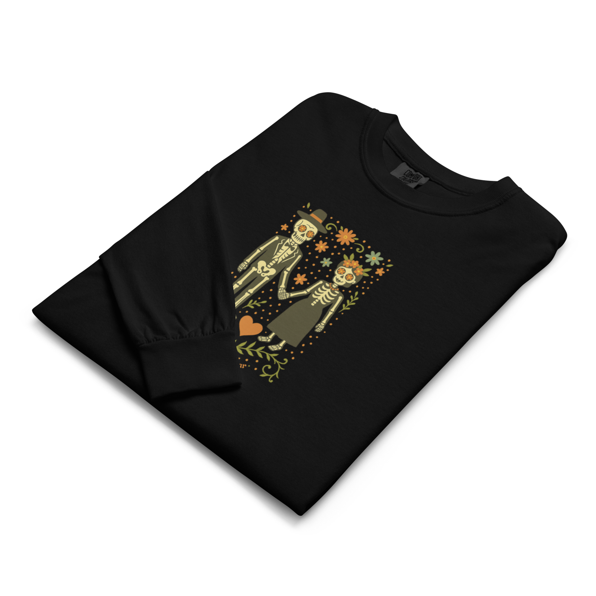 This is a folded black long sleeve shirt with a Día de los Muertos–style illustration of a male and female skeleton holding hands, surrounded by marigolds, vines, and folk art patterns. The skeletons wear traditional black clothing, the man in a suit and hat, the woman with a floral crown with a red heart and leaves at the bottom and the text Creepy All Year. Rendered in warm tones of orange, green, and cream, symbolizing eternal love and remembrance
