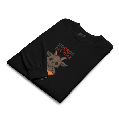 This is a folded zoom in of a black long sleeve shirt with a cartoon illustration of a baby Baphomet goat with curved horns and a playful grin, wearing bright floral swim trunks while floating in a colorful inner tube. The character is set against a light background with the bold text “Horror All Year” above in red letters
