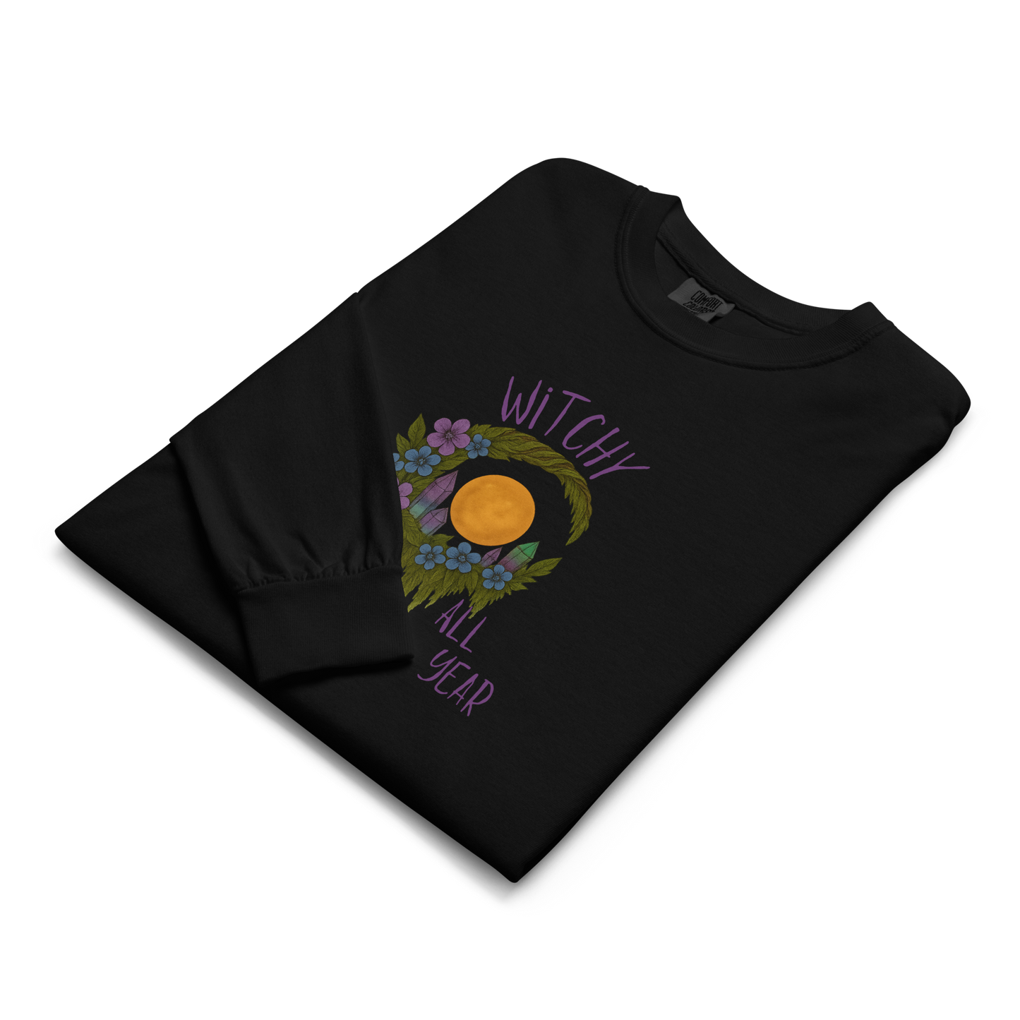 This is a folded black long sleeve shirt with a design of a hand-drawn wreath made of moss-covered branches wraps around a glowing sun. The wreath is decorated with purple and blue flowers, small amethyst and fluorite crystals, and strands of hanging moss. The words “Witchy” curve above the wreath and “All Year” sits on the bottom right 