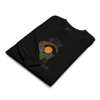 This is a folded black long sleeve shirt with a design of a hand-drawn wreath made of moss-covered branches wraps around a glowing sun. The wreath is decorated with purple and blue flowers, small amethyst and fluorite crystals, and strands of hanging moss. The words “Witchy” curve above the wreath and “All Year” sits on the bottom right 