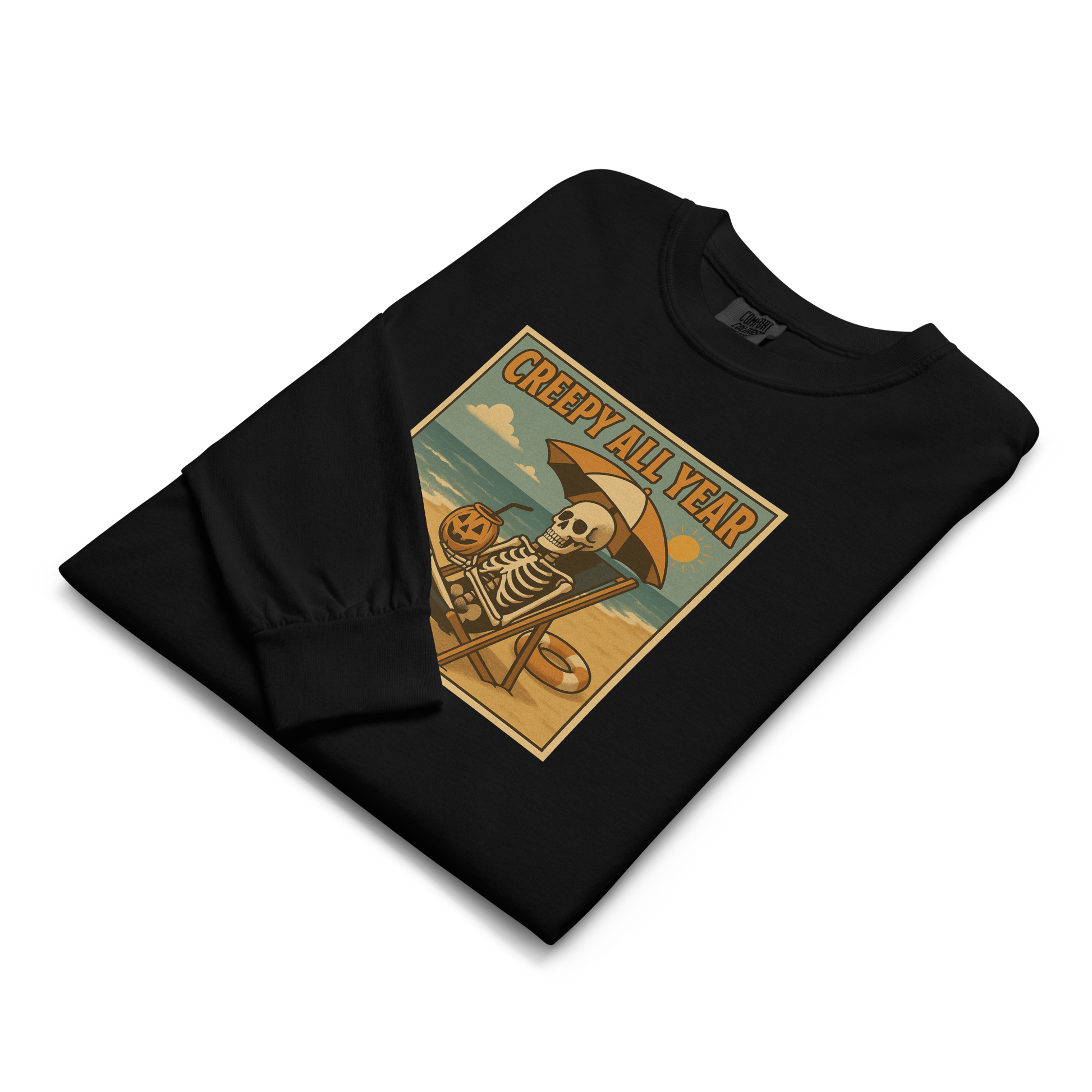 This is a folded black long-sleeve shirt with a retro-style illustration of a grinning skeleton lounging on a beach chair under an orange umbrella. The skeleton is holding a pumpkin-shaped drink cup with a straw and relaxing by the ocean, with a bright sun in the sky and a life preserver in the sand. The words “Creepy All Year” appear boldly at the top