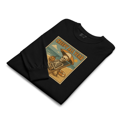 This is a folded black long-sleeve shirt with a retro-style illustration of a grinning skeleton lounging on a beach chair under an orange umbrella. The skeleton is holding a pumpkin-shaped drink cup with a straw and relaxing by the ocean, with a bright sun in the sky and a life preserver in the sand. The words “Creepy All Year” appear boldly at the top