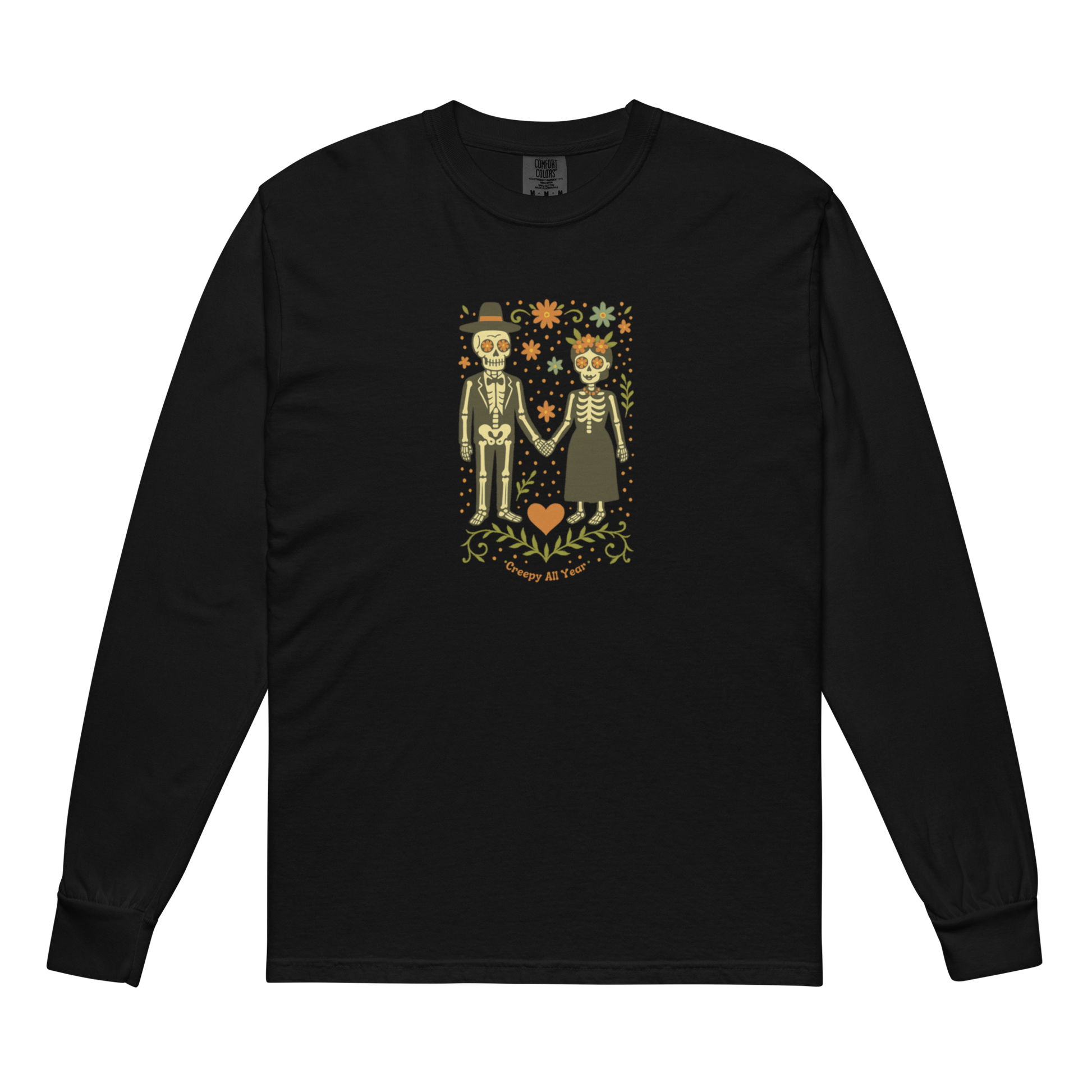 This is a black long sleeve shirt with a Día de los Muertos–style illustration of a male and female skeleton holding hands, surrounded by marigolds, vines, and folk art patterns. The skeletons wear traditional black clothing, the man in a suit and hat, the woman with a floral crown with a red heart and leaves at the bottom and the text Creepy All Year. Rendered in warm tones of orange, green, and cream, symbolizing eternal love and remembrance