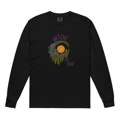 This is a black long sleeve shirt with a design of a hand-drawn wreath made of moss-covered branches wraps around a glowing sun. The wreath is decorated with purple and blue flowers, small amethyst and fluorite crystals, and strands of hanging moss. The words “Witchy” curve above the wreath and “All Year” sits on the bottom right 