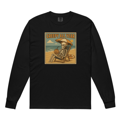 This is a black long-sleeve shirt with a retro-style illustration of a grinning skeleton lounging on a beach chair under an orange umbrella. The skeleton is holding a pumpkin-shaped drink cup with a straw and relaxing by the ocean, with a bright sun in the sky and a life preserver in the sand. The words “Creepy All Year” appear boldly at the top