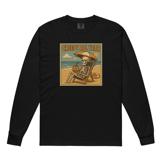 This is a black long-sleeve shirt with a retro-style illustration of a grinning skeleton lounging on a beach chair under an orange umbrella. The skeleton is holding a pumpkin-shaped drink cup with a straw and relaxing by the ocean, with a bright sun in the sky and a life preserver in the sand. The words “Creepy All Year” appear boldly at the top