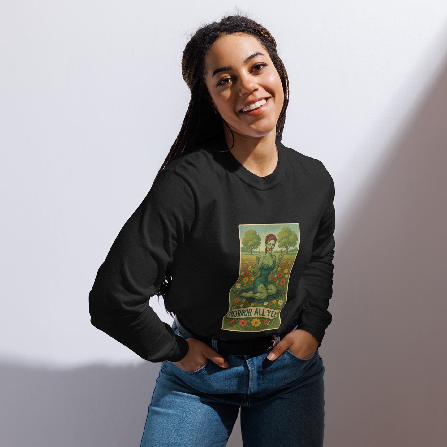 This is a woman with dark braids wearing a black long sleeve shirt with a design illustration of a sassy zombie pinup girl standing in a floral field, wearing tattered lingerie and flipping off the viewer with both hands. She has green skin, vintage curls, and a bold attitude. The design blends horror and humor, with a pastel color palette and creepy cute vibes