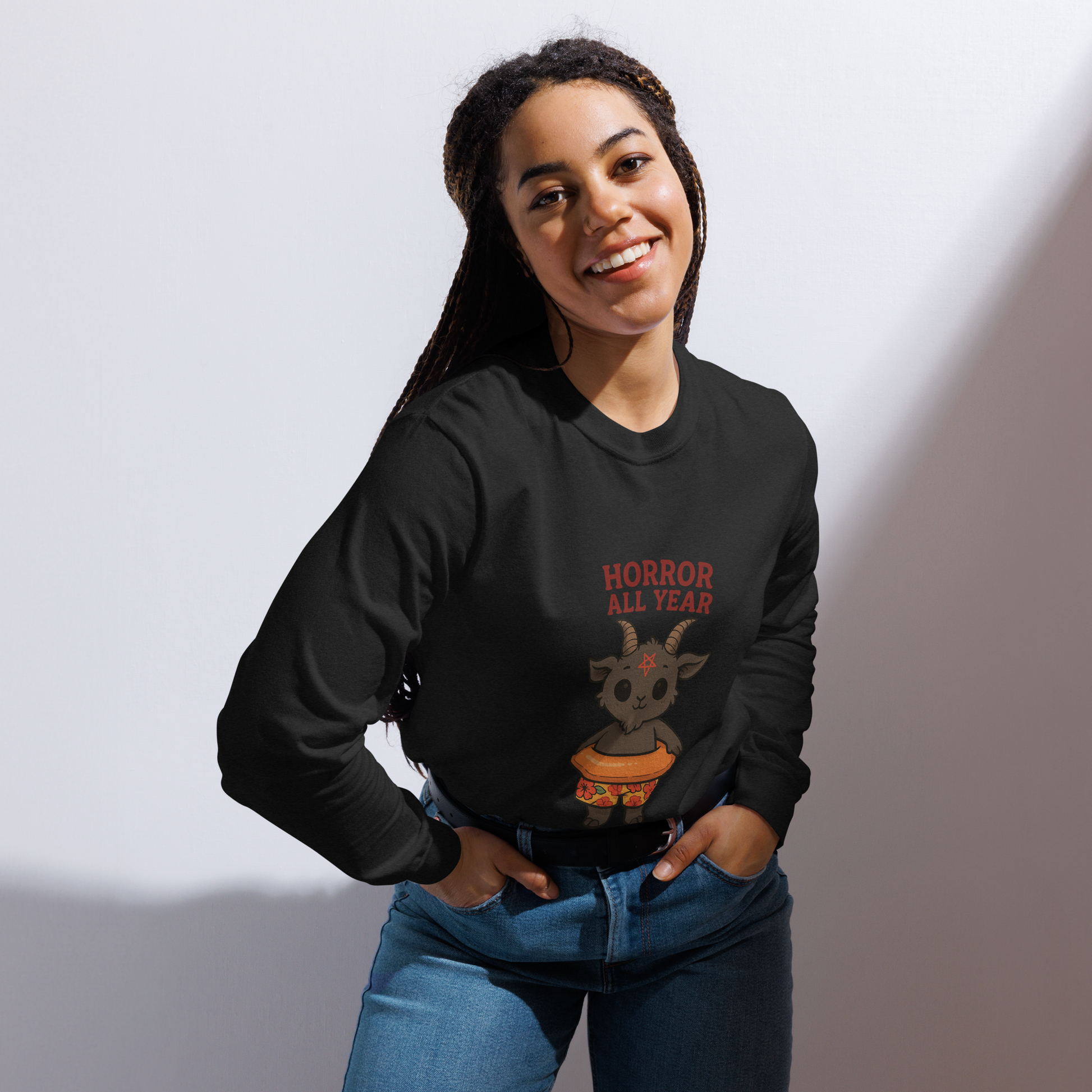 This is a woman with dark braided hair wearing a black long sleeve shirt with a cartoon illustration of a baby Baphomet goat with curved horns and a playful grin, wearing bright floral swim trunks while floating in a colorful inner tube. The character is set against a light background with the bold text “Horror All Year” above in red letters
