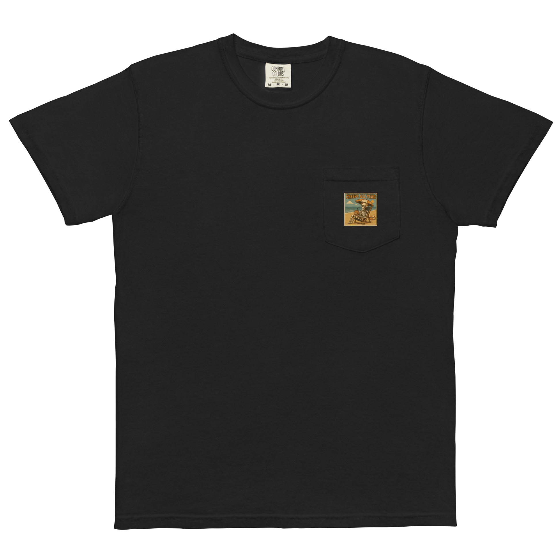 This is a black pocket t-shirt with a retro-style illustration of a grinning skeleton lounging on a beach chair under an orange umbrella. The skeleton is holding a pumpkin-shaped drink cup with a straw and relaxing by the ocean, with a bright sun in the sky and a life preserver in the sand. The words “Creepy All Year” appear boldly at the top