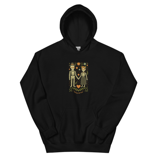 This is a black hoodie with a design of a Día de los Muertos–style illustration of a male and female skeleton holding hands, surrounded by marigolds, vines, and folk art patterns. The skeletons wear traditional black clothing, the man in a suit and hat, the woman with a floral crown with a red heart and leaves at the bottom and the text Creepy All Year. Rendered in warm tones of orange, green, and cream, symbolizing eternal love and remembrance