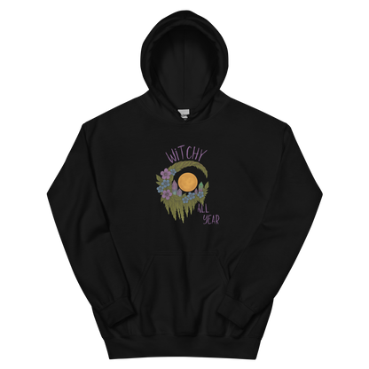 This is a black hoodie with a design of a hand-drawn wreath made of moss-covered branches wraps around a glowing sun. The wreath is decorated with purple and blue flowers, small amethyst and fluorite crystals, and strands of hanging moss. The words “Witchy” curve above the wreath and “All Year” sits on the bottom right