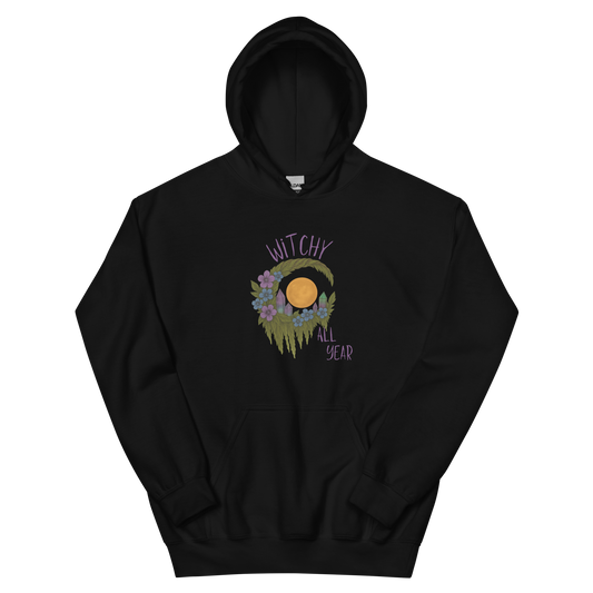 This is a black hoodie with a design of a hand-drawn wreath made of moss-covered branches wraps around a glowing sun. The wreath is decorated with purple and blue flowers, small amethyst and fluorite crystals, and strands of hanging moss. The words “Witchy” curve above the wreath and “All Year” sits on the bottom right