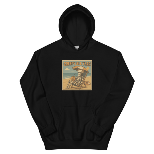 This is a black hoodie with a retro-style illustration of a grinning skeleton lounging on a beach chair under an orange umbrella. The skeleton is holding a pumpkin-shaped drink cup with a straw and relaxing by the ocean, with a bright sun in the sky and a life preserver in the sand. The words “Creepy All Year” appear boldly at the top