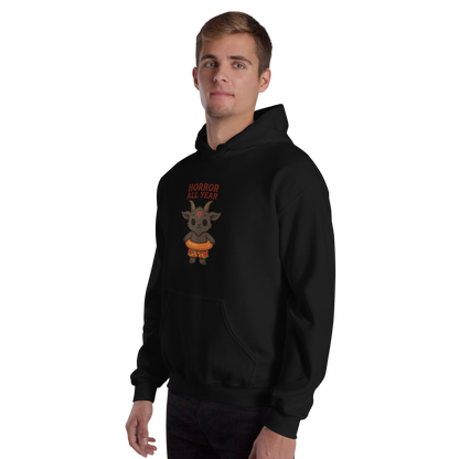 This is a man with light hair wearing a black hoodie with a cartoon illustration of a baby Baphomet goat with curved horns and a playful grin, wearing bright floral swim trunks while floating in a colorful inner tube. The character is set against a light background with the bold text “Horror All Year” above in red letters