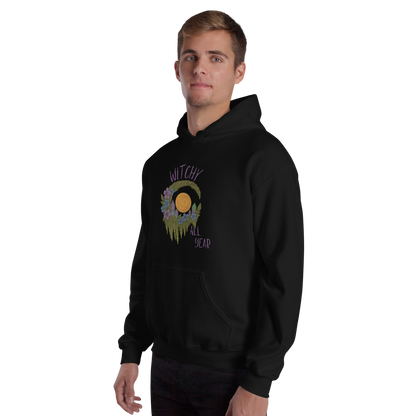 This is a man with blonde hair wearing a black hoodie with a design of a hand-drawn wreath made of moss-covered branches wraps around a glowing sun. The wreath is decorated with purple and blue flowers, small amethyst and fluorite crystals, and strands of hanging moss. The words “Witchy” curve above the wreath and “All Year” sits on the bottom right