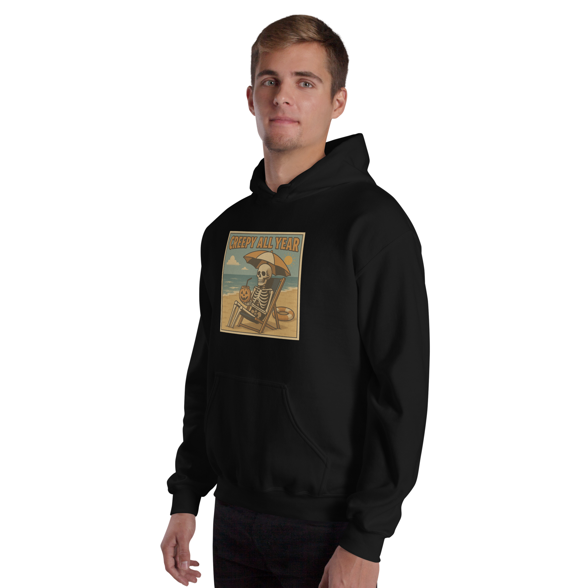 This is a man with light hair wearing black hoodie with a retro-style illustration of a grinning skeleton lounging on a beach chair under an orange umbrella. The skeleton is holding a pumpkin-shaped drink cup with a straw and relaxing by the ocean, with a bright sun in the sky and a life preserver in the sand. The words “Creepy All Year” appear boldly at the top 