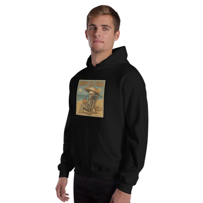 This is a man with light hair wearing black hoodie with a retro-style illustration of a grinning skeleton lounging on a beach chair under an orange umbrella. The skeleton is holding a pumpkin-shaped drink cup with a straw and relaxing by the ocean, with a bright sun in the sky and a life preserver in the sand. The words “Creepy All Year” appear boldly at the top 
