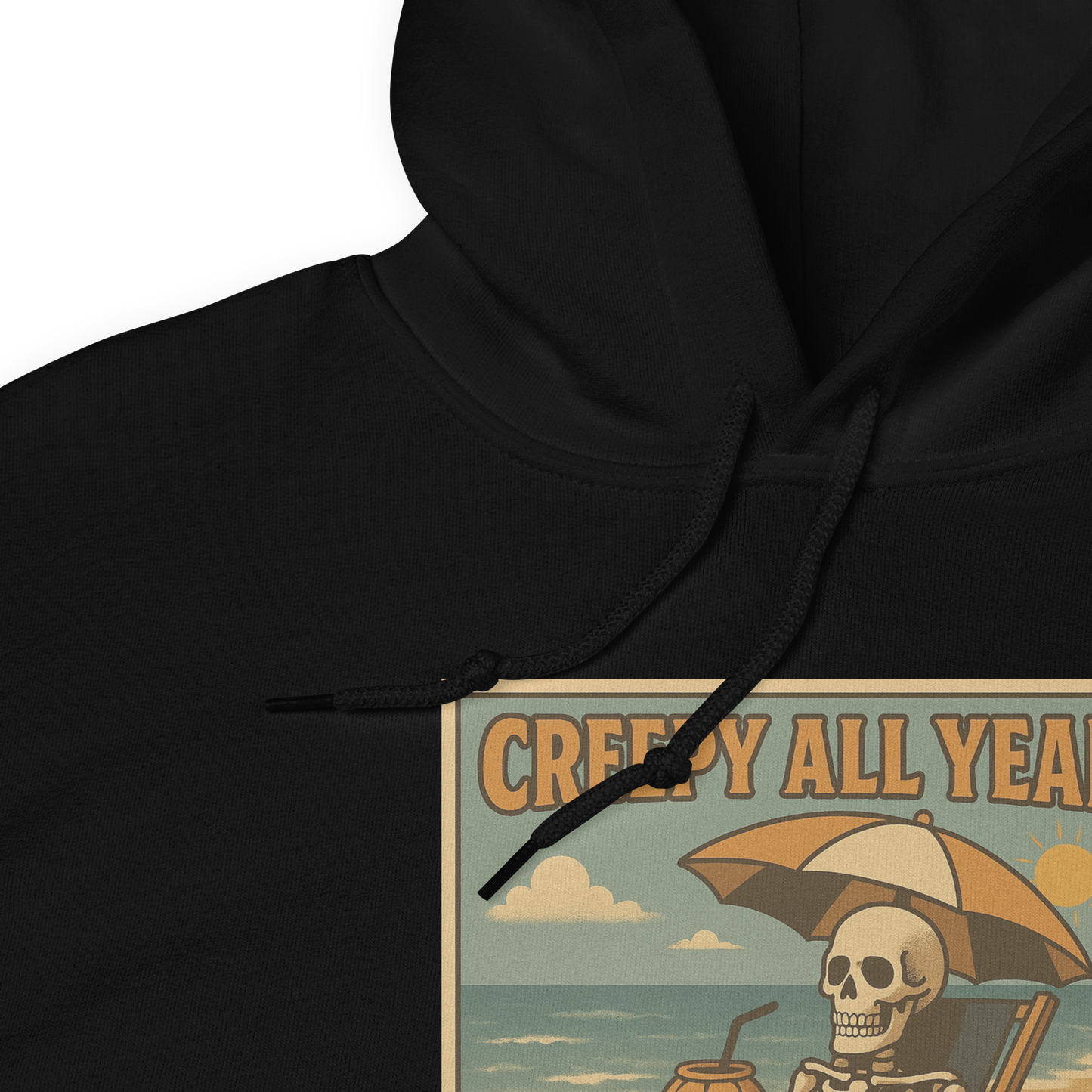 This is a zoomed in black hoodie with a retro-style illustration of a grinning skeleton lounging on a beach chair under an orange umbrella. The skeleton is holding a pumpkin-shaped drink cup with a straw and relaxing by the ocean, with a bright sun in the sky and a life preserver in the sand. The words “Creepy All Year” appear boldly at the top 