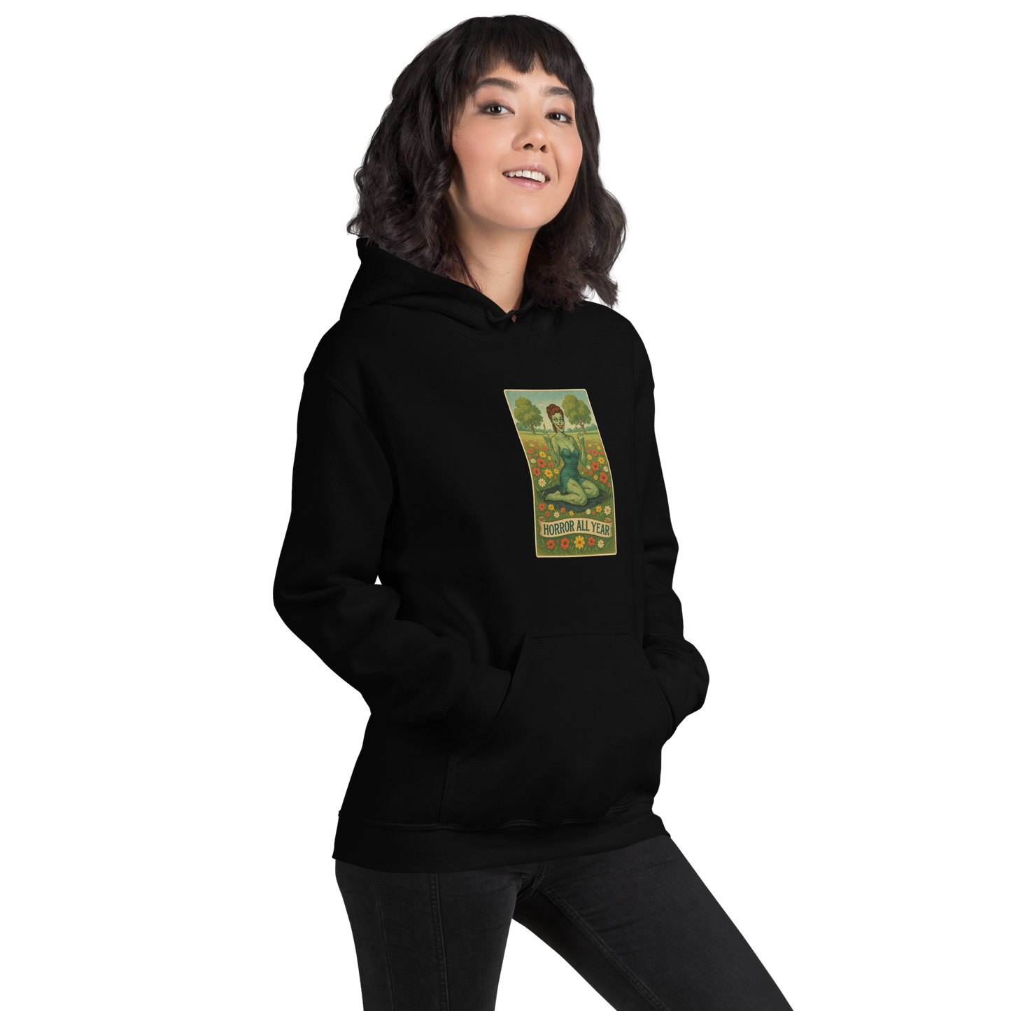 This is a woman with dark hair wearing a black hoodie with a design illustration of a sassy zombie pinup girl standing in a floral field, wearing tattered lingerie and flipping off the viewer with both hands. She has green skin, vintage curls, and a bold attitude. The design blends horror and humor, with a pastel color palette and creepy cute vibes.