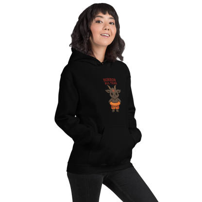 This is a girl with dark hair wearing a black hoodie with a cartoon illustration of a baby Baphomet goat with curved horns and a playful grin, wearing bright floral swim trunks while floating in a colorful inner tube. The character is set against a light background with the bold text “Horror All Year” above in red letters