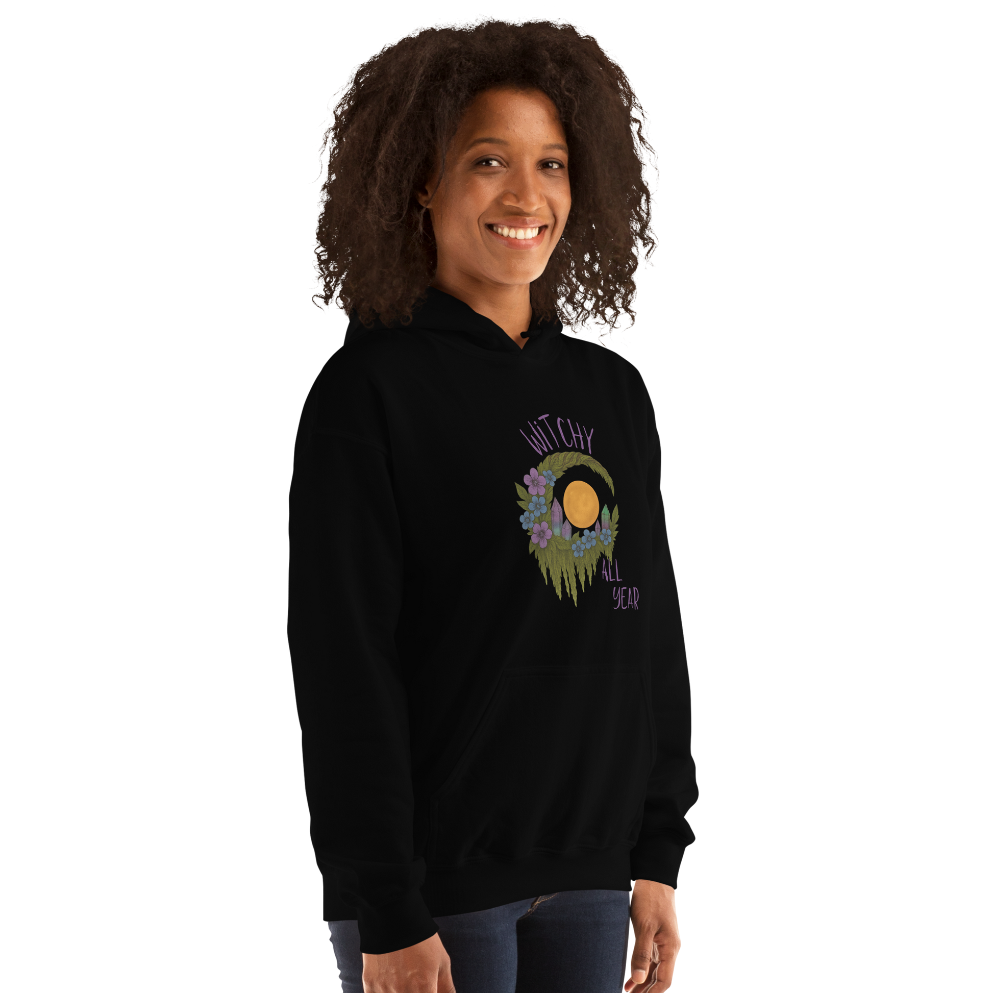 This is a woman with dark hair wearing a black hoodie with a design of a hand-drawn wreath made of moss-covered branches wraps around a glowing sun. The wreath is decorated with purple and blue flowers, small amethyst and fluorite crystals, and strands of hanging moss. The words “Witchy” curve above the wreath and “All Year” sits on the bottom right