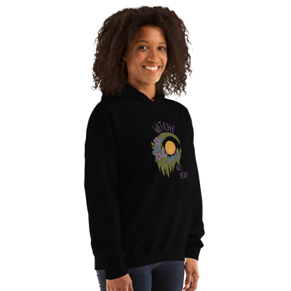 This is a woman with dark hair wearing a black hoodie with a design of a hand-drawn wreath made of moss-covered branches wraps around a glowing sun. The wreath is decorated with purple and blue flowers, small amethyst and fluorite crystals, and strands of hanging moss. The words “Witchy” curve above the wreath and “All Year” sits on the bottom right