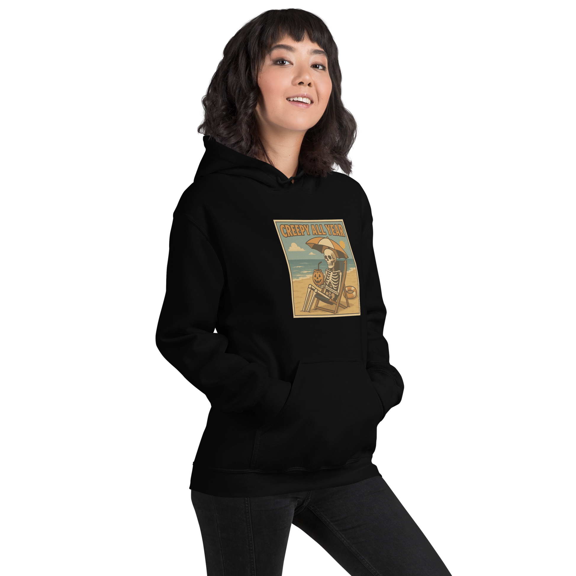 This is a woman with dark hair wearing black hoodie with a retro-style illustration of a grinning skeleton lounging on a beach chair under an orange umbrella. The skeleton is holding a pumpkin-shaped drink cup with a straw and relaxing by the ocean, with a bright sun in the sky and a life preserver in the sand. The words “Creepy All Year” appear boldly at the top 