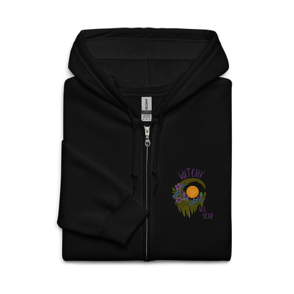 This is a folded black zip-up hoodie with a design of a hand-drawn wreath made of moss-covered branches wraps around a glowing sun. The wreath is decorated with purple and blue flowers, small amethyst and fluorite crystals, and strands of hanging moss. The words “Witchy” curve above the wreath and “All Year” sits on the bottom right