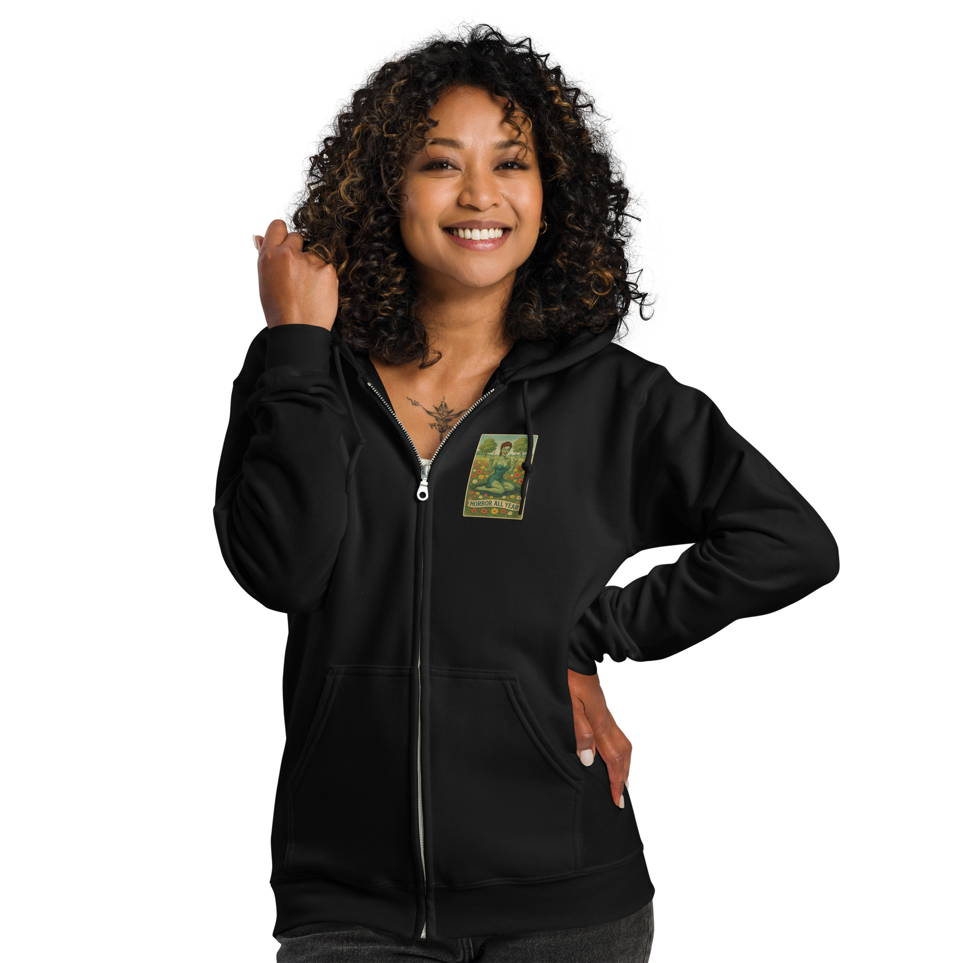 This is a woman with dark curly hair wearing a black zip-up hoodie with a design illustration of a sassy zombie pinup girl standing in a floral field, wearing tattered lingerie and flipping off the viewer with both hands. She has green skin, vintage curls, and a bold attitude. The design blends horror and humor, with a pastel color palette and creepy cute vibes.