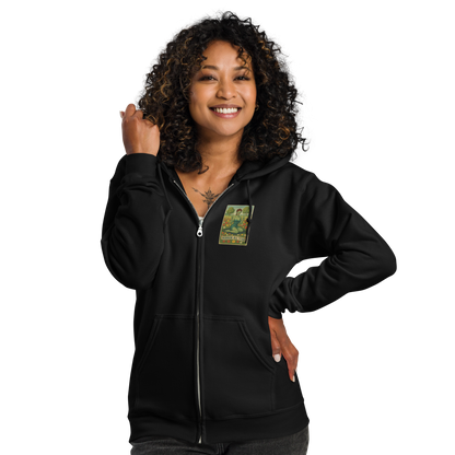 This is a woman with dark curly hair wearing a black zip-up hoodie with a design illustration of a sassy zombie pinup girl standing in a floral field, wearing tattered lingerie and flipping off the viewer with both hands. She has green skin, vintage curls, and a bold attitude. The design blends horror and humor, with a pastel color palette and creepy cute vibes.