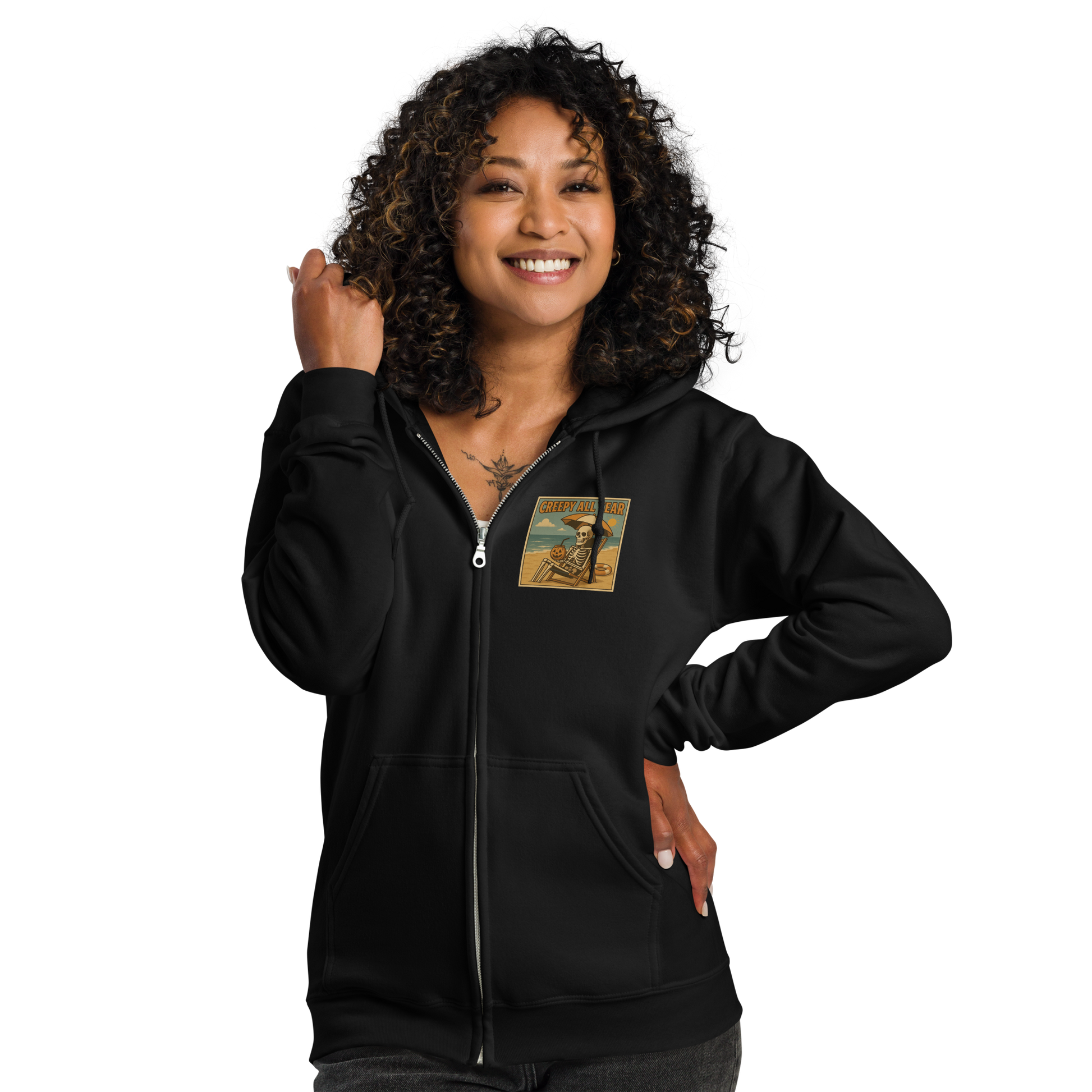 This is a woman with black curly hair wearing a black zip-up hoodie with a retro-style illustration of a grinning skeleton lounging on a beach chair under an orange umbrella. The skeleton is holding a pumpkin-shaped drink cup with a straw and relaxing by the ocean, with a bright sun in the sky and a life preserver in the sand. The words “Creepy All Year” appear boldly at the top