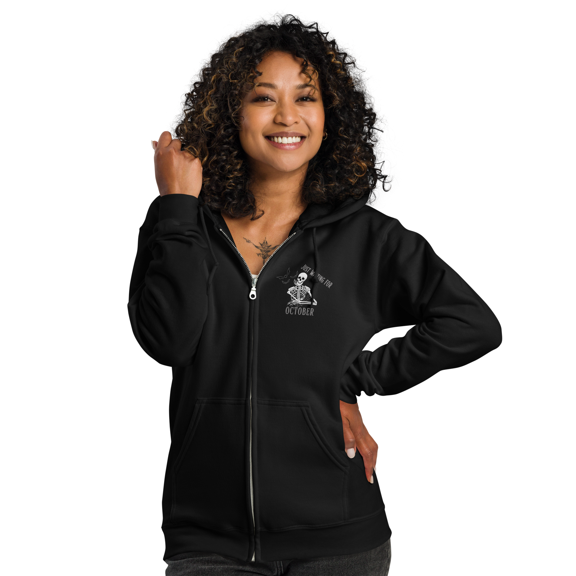 This is a woman with dark hair wearing a black zip-up hoodie that has a bored skeleton with his head resting in his hand and he has spider webs and bats around him and the text reads just waiting for October