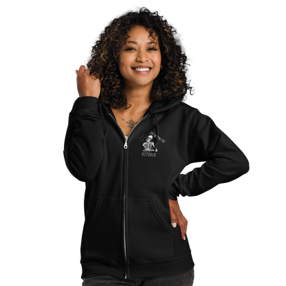 This is a woman with dark hair wearing a black zip-up hoodie that has a bored skeleton with his head resting in his hand and he has spider webs and bats around him and the text reads just waiting for October