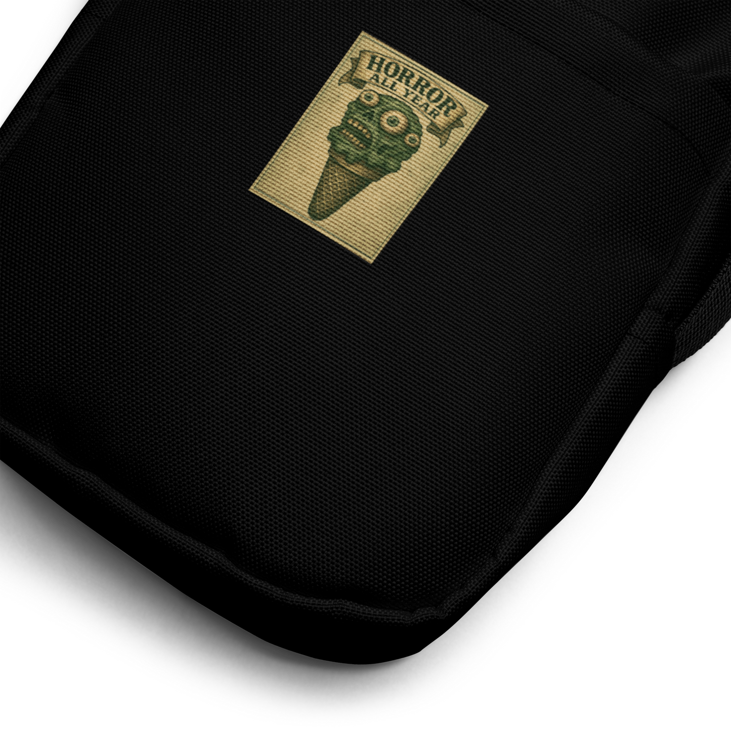 This is a zoom in of a black crossbody bag that is a vintage-style illustration of a zombie-themed ice cream cone with a green, melting undead face and bulging eyeballs. Above the cone, a horror banner reads "Horror All Year" on a distressed parchment background