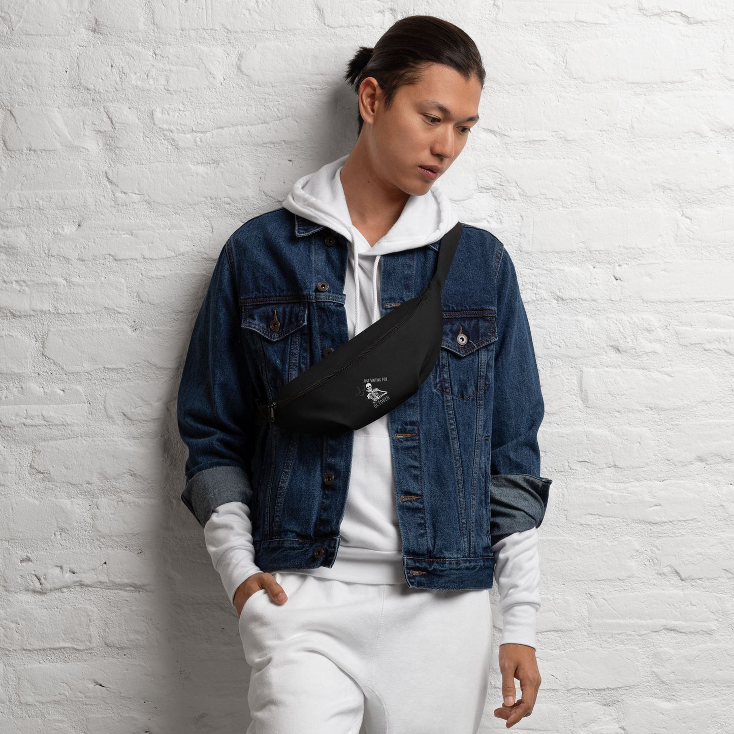 This is a man with dark hair wearing a black waist bag fanny pack that has a bored skeleton with his head resting in his hand and he has spider webs and bats around him and the text reads just waiting for October