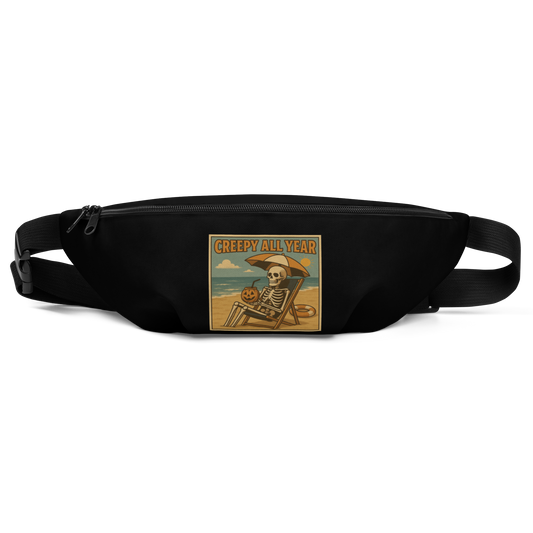 This is a black waist bag fanny pack with a retro-style illustration of a grinning skeleton lounging on a beach chair under an orange umbrella. The skeleton is holding a pumpkin-shaped drink cup with a straw and relaxing by the ocean, with a bright sun in the sky and a life preserver in the sand. The words “Creepy All Year” appear boldly at the top 