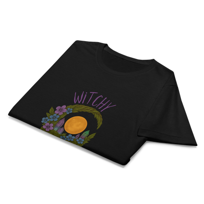 This is a folded black women's t-shirt with a design of a hand-drawn wreath made of moss-covered branches wraps around a glowing sun. The wreath is decorated with purple and blue flowers, small amethyst and fluorite crystals, and strands of hanging moss. The words “Witchy” curve above the wreath and “All Year” sits on the bottom right 
