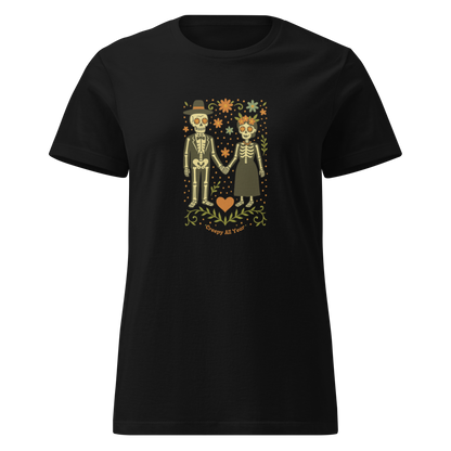 This is a black women's t-shirt with a Día de los Muertos–style illustration of a male and female skeleton holding hands, surrounded by marigolds, vines, and folk art patterns. The skeletons wear traditional black clothing, the man in a suit and hat, the woman with a floral crown with a red heart and leaves at the bottom and the text Creepy All Year. Rendered in warm tones of orange, green, and cream, symbolizing eternal love and remembrance
