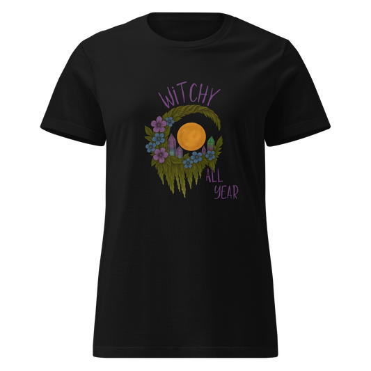 This a black women's t-shirt with a design of a hand-drawn wreath made of moss-covered branches wraps around a glowing sun. The wreath is decorated with purple and blue flowers, small amethyst and fluorite crystals, and strands of hanging moss. The words “Witchy” curve above the wreath and “All Year” sits on the bottom right 