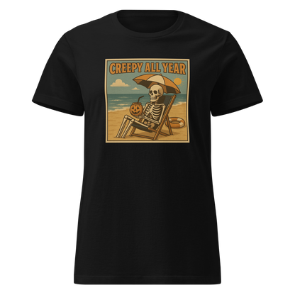 This is a black style woman's t-shirt with a retro-style illustration of a grinning skeleton lounging on a beach chair under an orange umbrella. The skeleton is holding a pumpkin-shaped drink cup with a straw and relaxing by the ocean, with a bright sun in the sky and a life preserver in the sand. The words “Creepy All Year” appear boldly at the top
