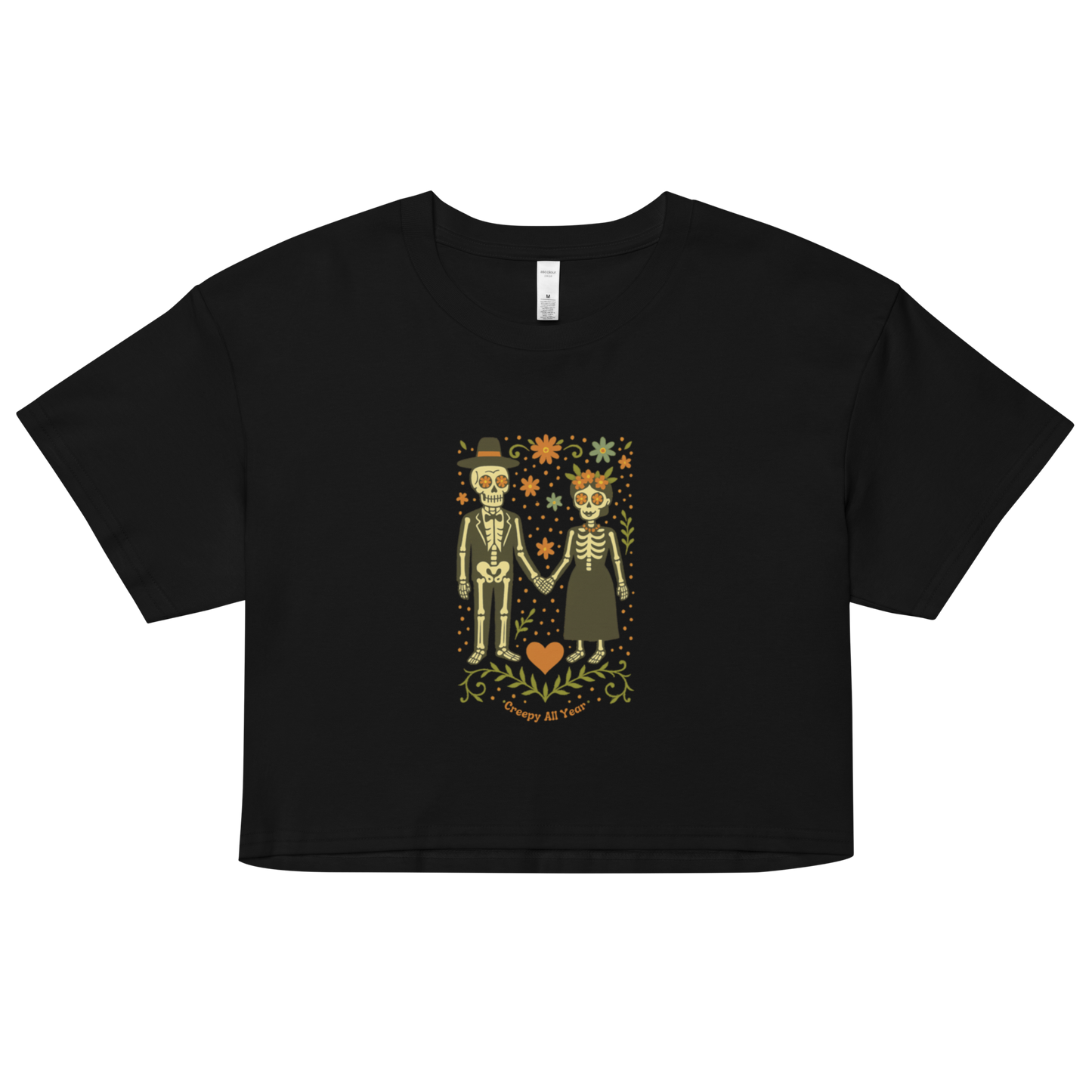 This is a black crop top with a Día de los Muertos–style illustration of a male and female skeleton holding hands, surrounded by marigolds, vines, and folk art patterns. The skeletons wear traditional black clothing, the man in a suit and hat, the woman with a floral crown with a red heart and leaves at the bottom and the text Creepy All Year. Rendered in warm tones of orange, green, and cream, symbolizing eternal love and remembrance 