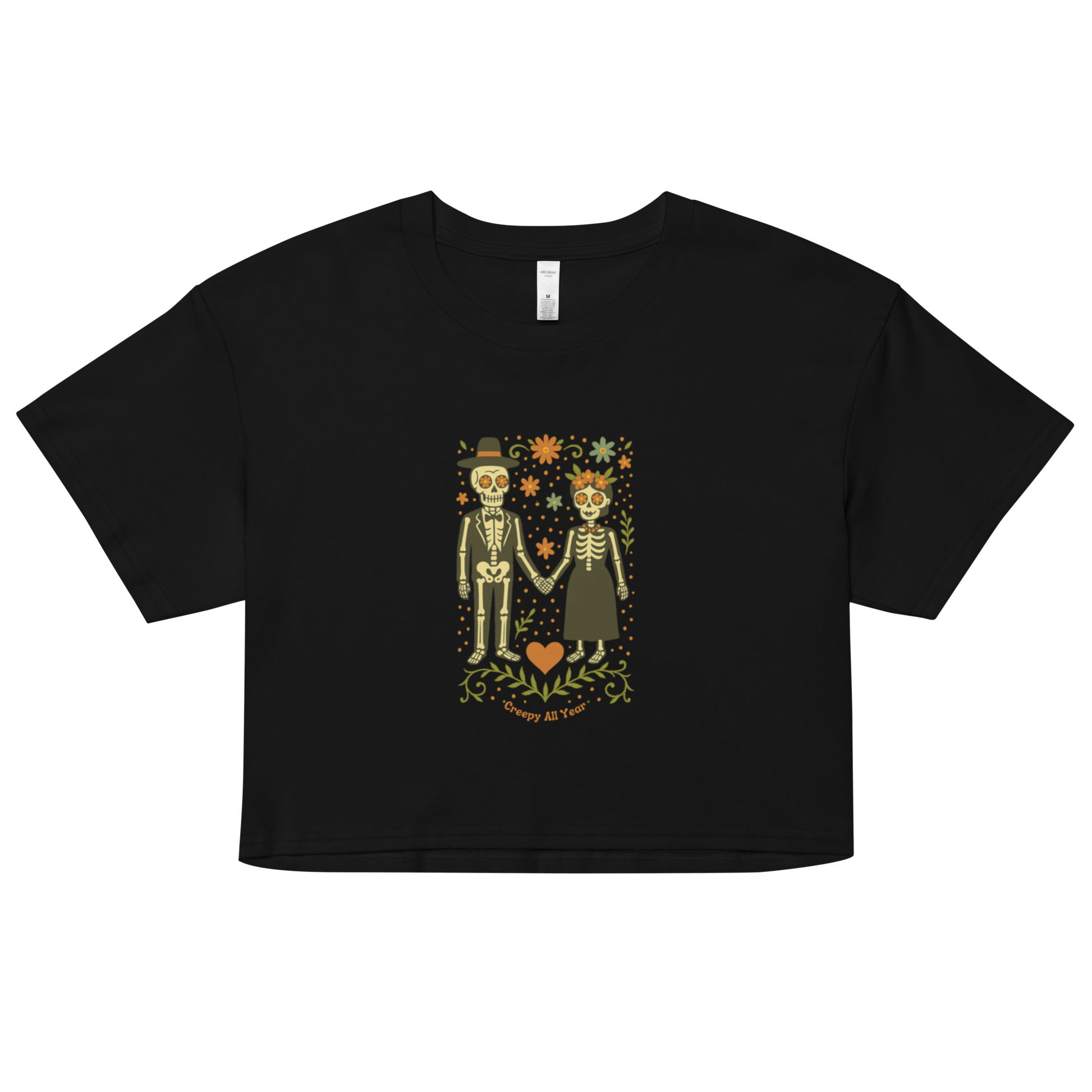 This is a black crop top with a Día de los Muertos–style illustration of a male and female skeleton holding hands, surrounded by marigolds, vines, and folk art patterns. The skeletons wear traditional black clothing, the man in a suit and hat, the woman with a floral crown with a red heart and leaves at the bottom and the text Creepy All Year. Rendered in warm tones of orange, green, and cream, symbolizing eternal love and remembrance 