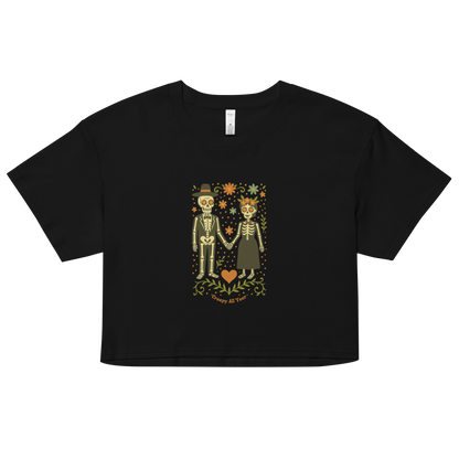 This is a black crop top with a Día de los Muertos–style illustration of a male and female skeleton holding hands, surrounded by marigolds, vines, and folk art patterns. The skeletons wear traditional black clothing, the man in a suit and hat, the woman with a floral crown with a red heart and leaves at the bottom and the text Creepy All Year. Rendered in warm tones of orange, green, and cream, symbolizing eternal love and remembrance 
